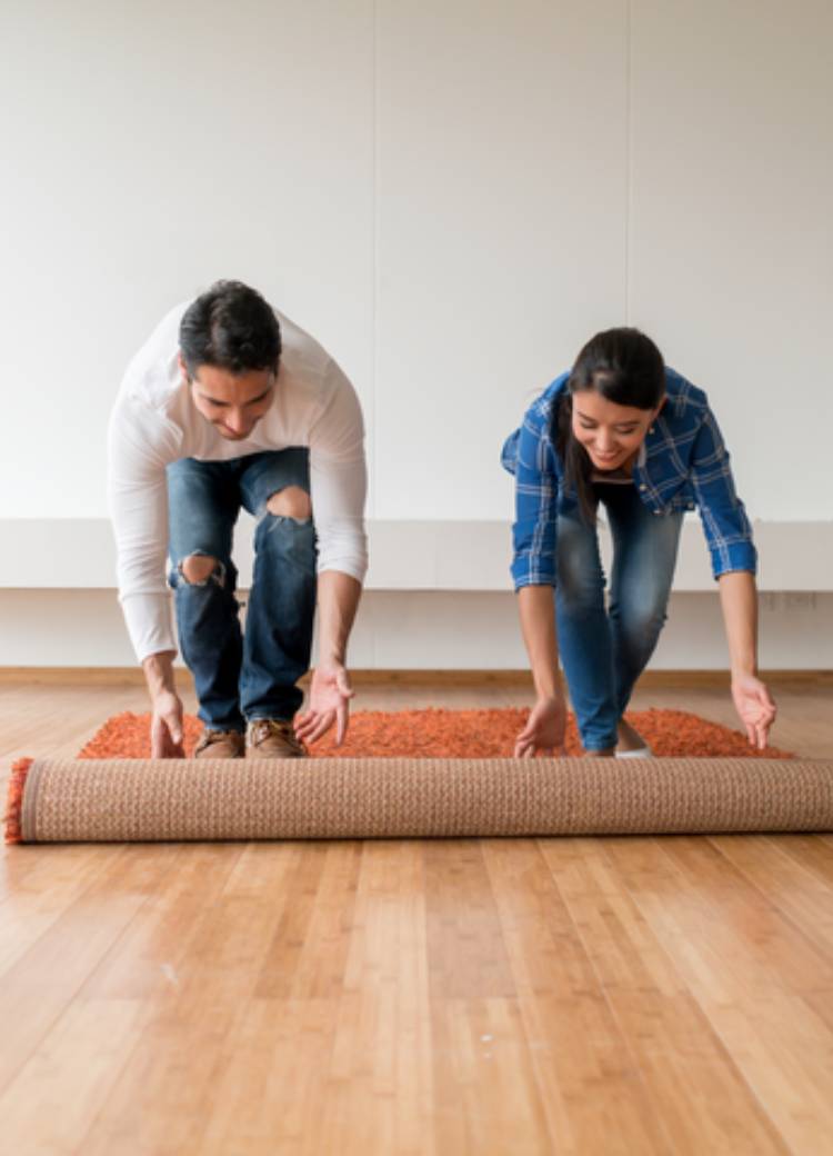 two people unrolling orange carpet in room with wood looking flooring and large windows.